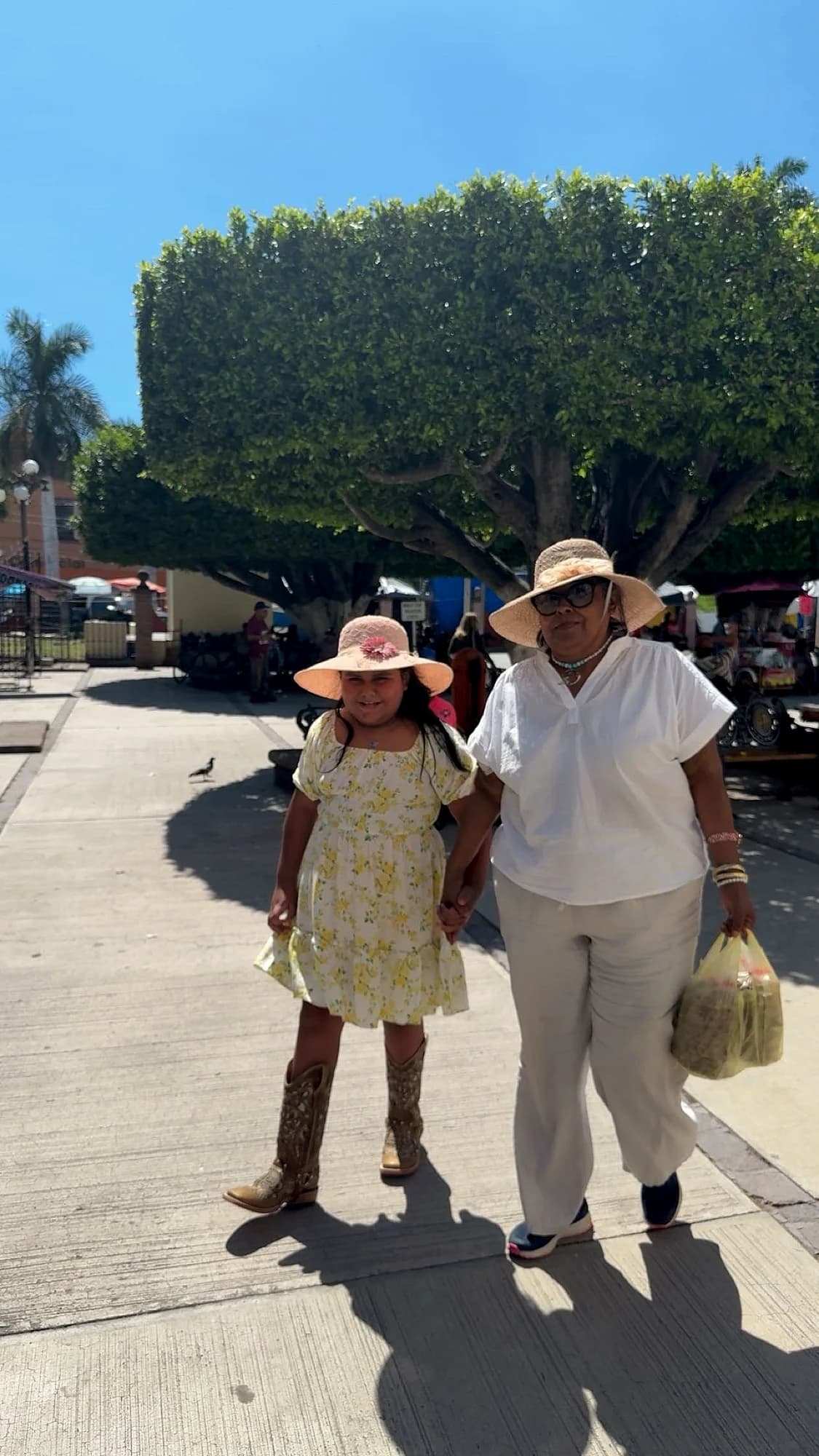 Maria shops at an outdoor market in Mexico, browsing items at a vendor's stall with her granddaughter.